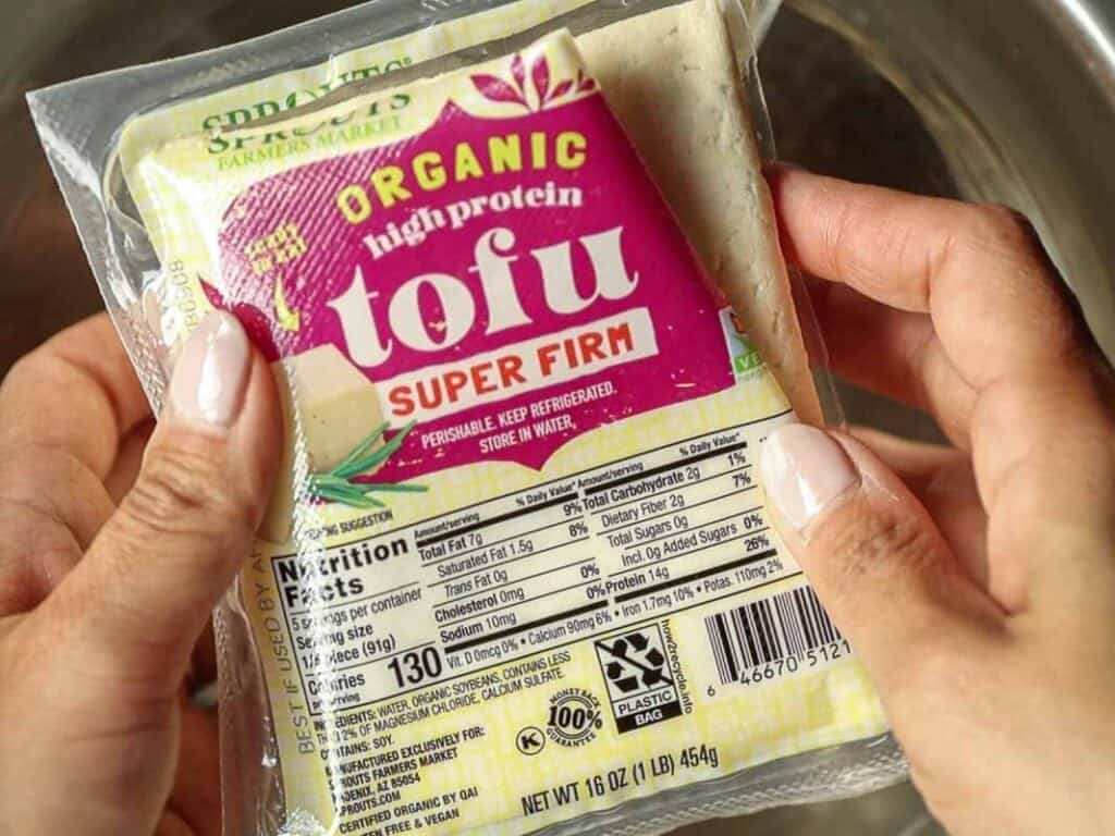 Close-up of hands holding a package of organic, high-protein, super firm tofu. The label shows it as non-GMO, with nutrition facts visible. The package weighs 16 oz (454g).